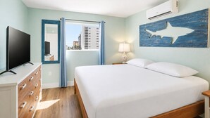 Ein Hotelzimmer mit einem Bett, einem Fernseher, einem Schminktisch, einem Fenster mit Vorhängen und einer Wandklimaanlage.