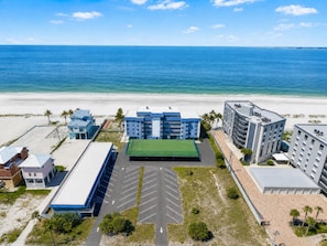Un hotel frente a la playa con varias construcciones, una cancha de tenis verde y un estacionamiento.