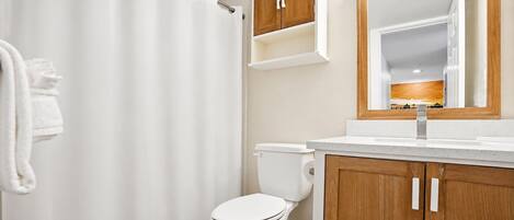 Un baño con cortina de ducha, inodoro, armarios de madera y un espejo sobre el lavamanos.