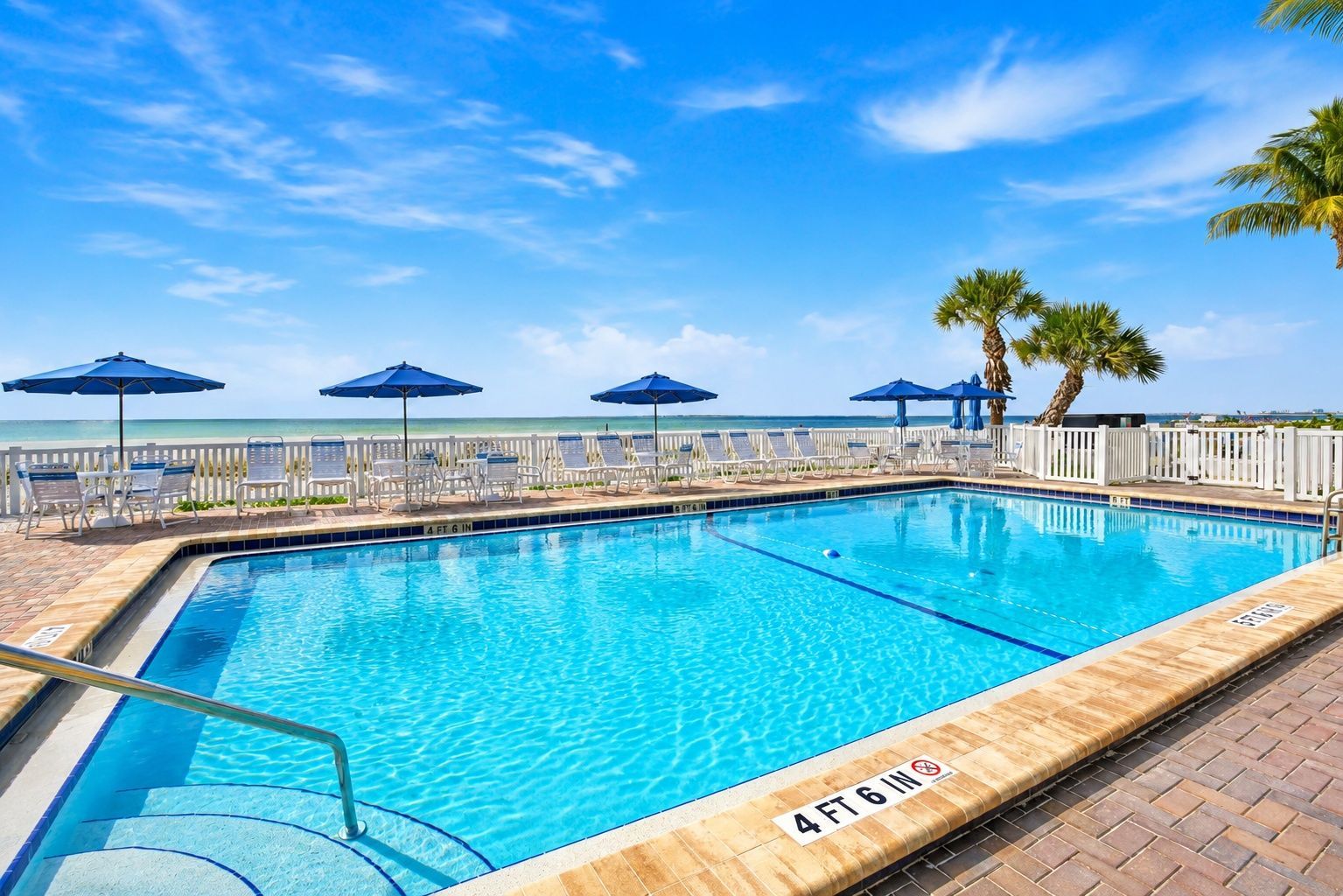 A rectangular swimming pool with a 4 ft 6 in depth marker, surrounded by lounge chairs and umbrellas, with a white fence and palm trees in the background.