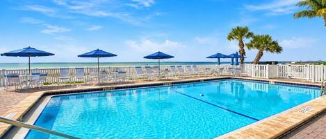 Piscina rettangolare con indicatore di profondità a 1,4 metri, circondata da sdraio e ombrelloni, con recinzione bianca e palme sullo sfondo.
