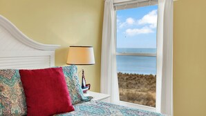 A bedroom with a bed, a nightstand with a lamp, and a window with a view of the sea.