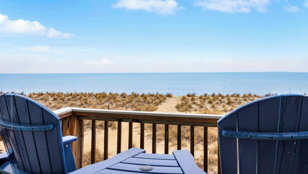 A beach view from a deck with two Adirondack chairs and a table, overlooking the ocean and dunes.