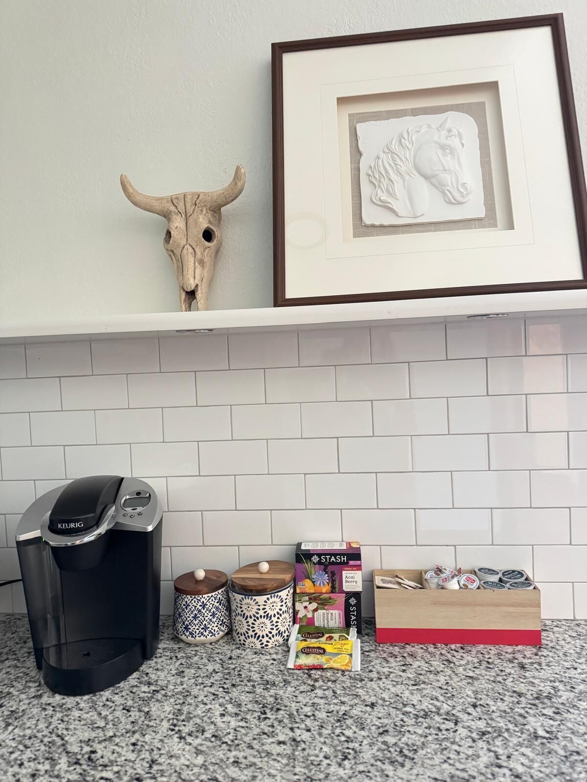 A kitchen counter with a Keurig coffee maker, two patterned containers, a framed picture of a bull, and a box of Stash tea bags.