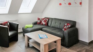 Ein modernes Wohnzimmer mit einem grauen Sofa, einem Holztisch und einem schwarzen Sessel. Zwei Fenster mit weißen Rahmen und ein Dachfenster sind vorhanden.