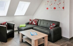 Ein modernes Wohnzimmer mit einem grauen Sofa, einem Holztisch und einem schwarzen Sessel. Zwei Fenster mit weißen Rahmen und ein Dachfenster sind vorhanden.
