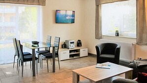 Una sala de estar moderna con una mesa de comedor de vidrio, sillas negras, un televisor de pantalla plana, una cafetera y una ventana con vistas al exterior.