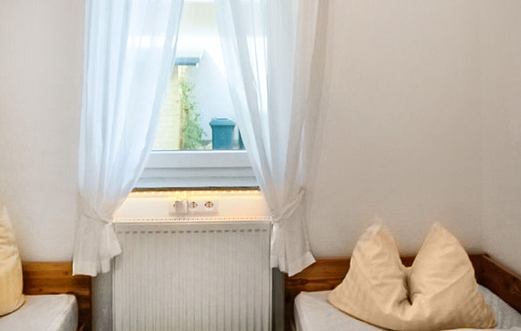 A bedroom with a window, white curtains, a radiator, and two pillows on the bed.