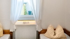 Un dormitorio con ventana, cortinas blancas, radiador y dos almohadas en la cama.