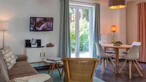 Un salon comprenant un canapé, une table à manger avec des chaises, un téléviseur fixé au mur et une fenêtre avec des rideaux.