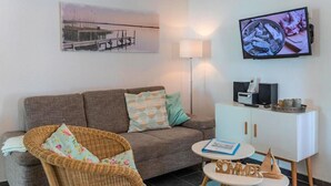 Un salon comprenant un canapé, un fauteuil en osier, une petite table avec un livre, un téléviseur fixé au mur et un meuble contenant des appareils électroniques.
