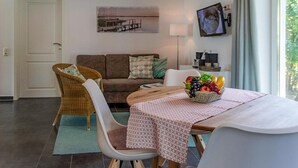 Un salon moderne avec un canapé, des fauteuils, une table à manger agrémentée d’un panier de fruits, une lampe et un téléviseur fixé au mur.