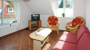 Un salón con un sofá rojo, dos sillones decorativos, un televisor sobre un mueble de madera y una ventana con vistas a un edificio de tejados rojos.