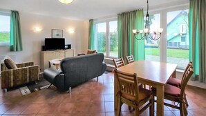 A dining area with a wooden table and chairs, a grey sofa, a television on a cabinet, and large windows with curtains. - 1 bedroom beautiful apartment in Röbel (Röbel)