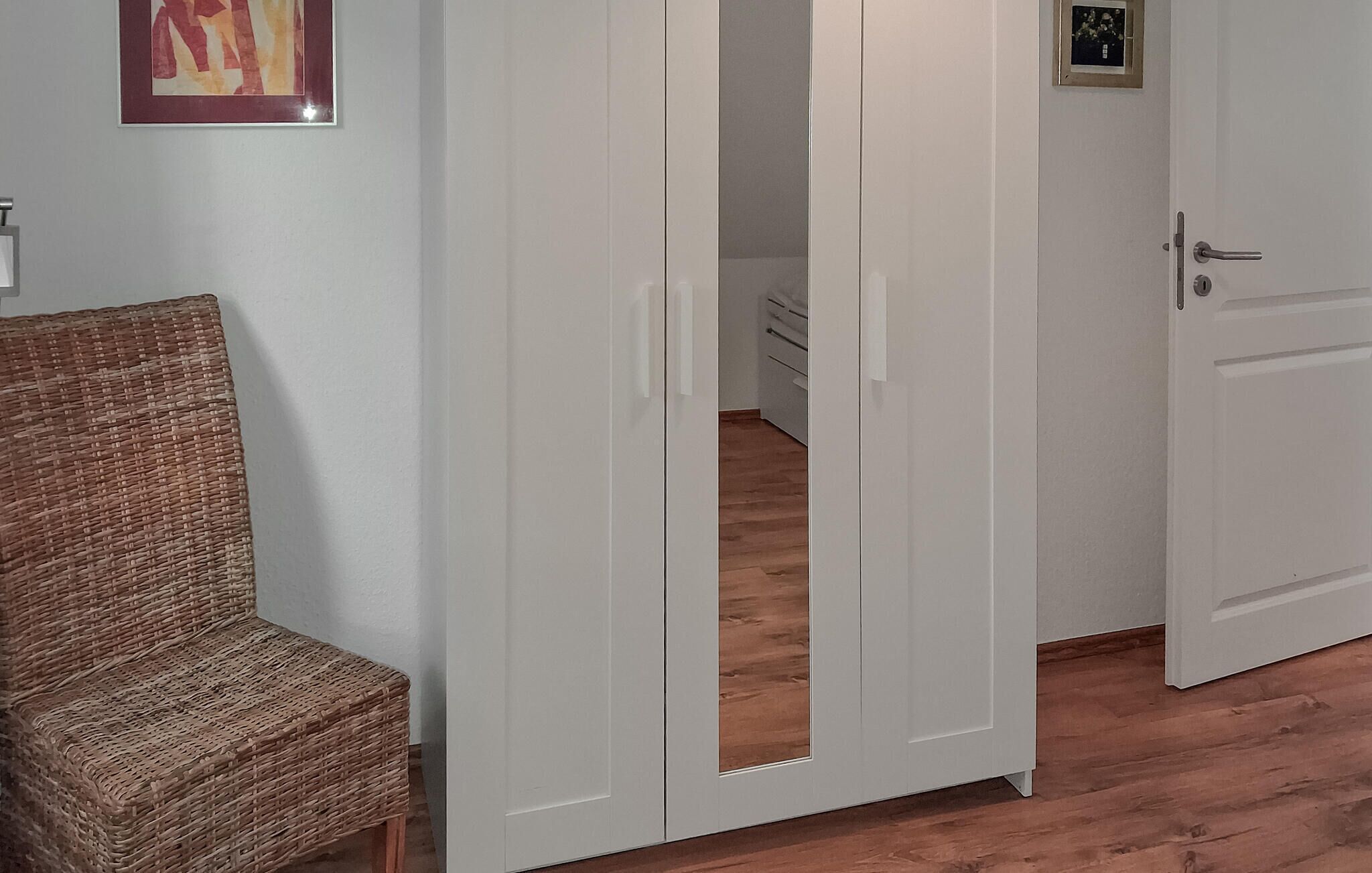 A room with a wicker chair, a white wardrobe, and a door leading to another room.