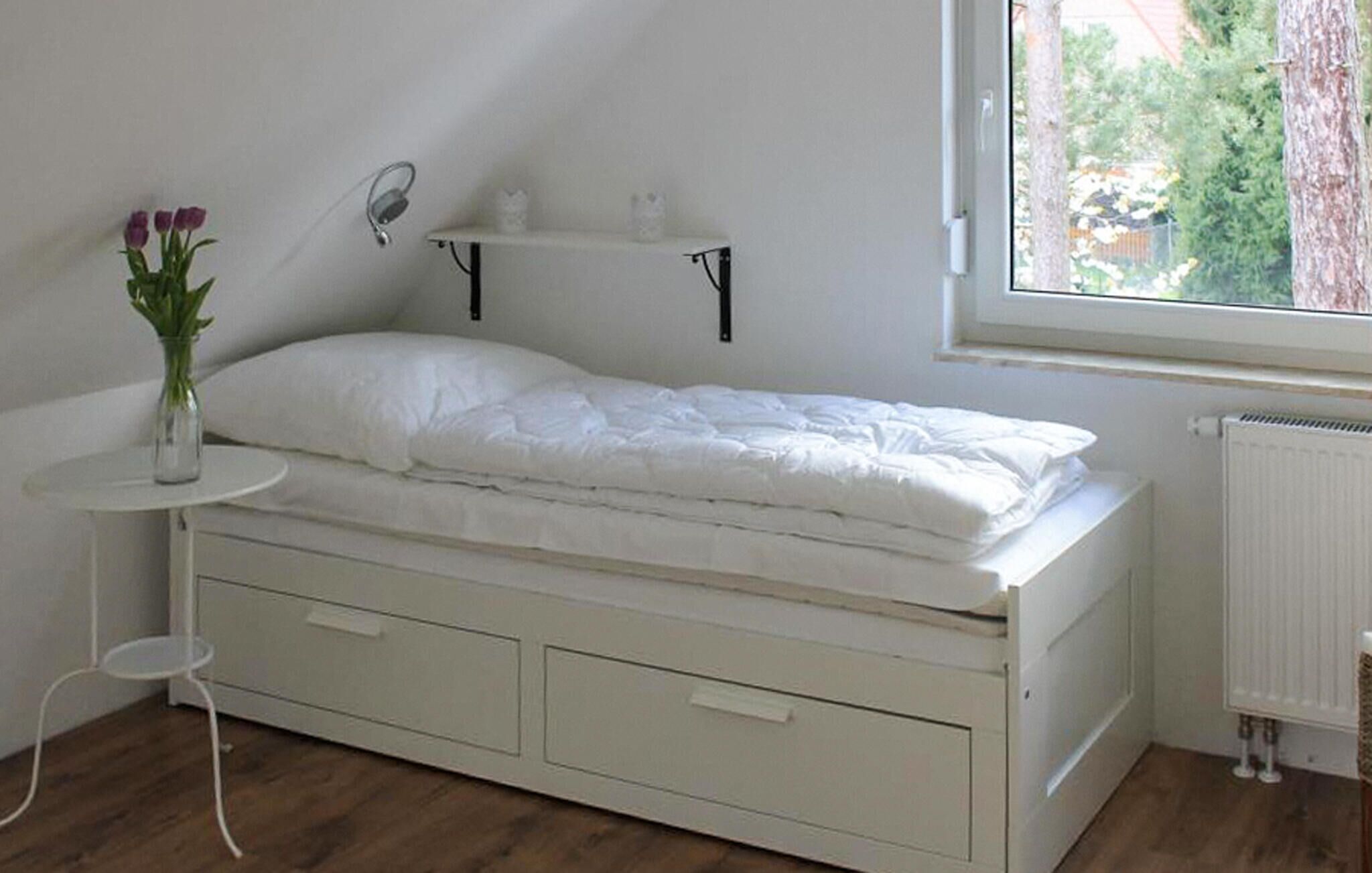 A small, white, single-bed room with a window, a radiator, and a small table with a vase of flowers.