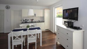 Eine kompakte Küche mit weißen Schränken, einem Esstisch mit Stühlen, einem Fernseher auf einer Kommode und einer Uhr an der Wand.