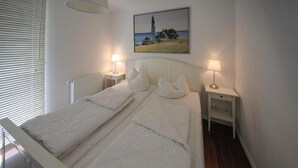Ein Doppelbett mit weißer Bettwäsche, zwei Nachttische mit Lampen, ein Gemälde eines Leuchtturms und ein Fenster mit Jalousien.