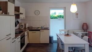 Eine moderne Küche mit weißen Schränken, einem eingebauten Backofen und einer Kaffeemaschine. Es gibt eine Essecke mit einem weißen Tisch und Stühlen, sowie ein Fenster mit Blick auf Grünflächen.