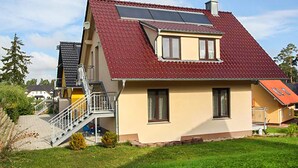 Una casa de dos pisos con techo rojo, un panel solar en el techo y una pequeña escalera que lleva a un balcón.