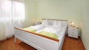 Habitación individual con una cama de madera, ropa de cama blanca, dos mesitas de noche con lámparas y una ventana con cortinas translúcidas.
