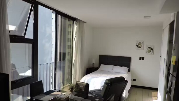 Quarto de hotel moderno com uma janela grande, uma cama com lençóis brancos, uma escrivaninha com um laptop e uma cadeira.