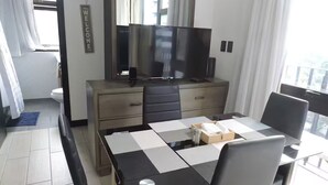 Quarto de hotel moderno com área de jantar, mesa com padrão xadrez preto e branco, TV de tela plana em móvel de madeira e vista da cidade através de grandes janelas.
