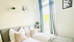 Ein ordentlich bezogenes Bett mit weißer und grauer Bettwäsche, ein Nachttisch mit Lampe und ein Fenster mit Vorhängen.