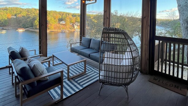 A wooden deck with a sofa, chairs, and a hanging chair overlooking a lake.