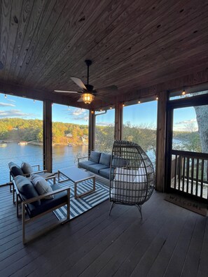 Terraza de madera con sofá, sillas y una sillona colgante con vista a un lago.