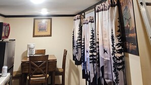 A rustic cabin interior with a deer head mounted on the ceiling, a dining area with wooden furniture, and forest-themed curtains.