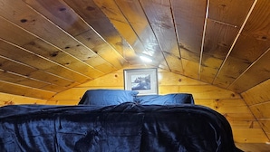 A cozy attic bedroom with wooden beams on the ceiling, a bed with a dark cover, and a framed picture on the wall.
