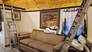 A cozy room with a wooden ceiling, a couch, a ladder, and a painting of a forest scene.