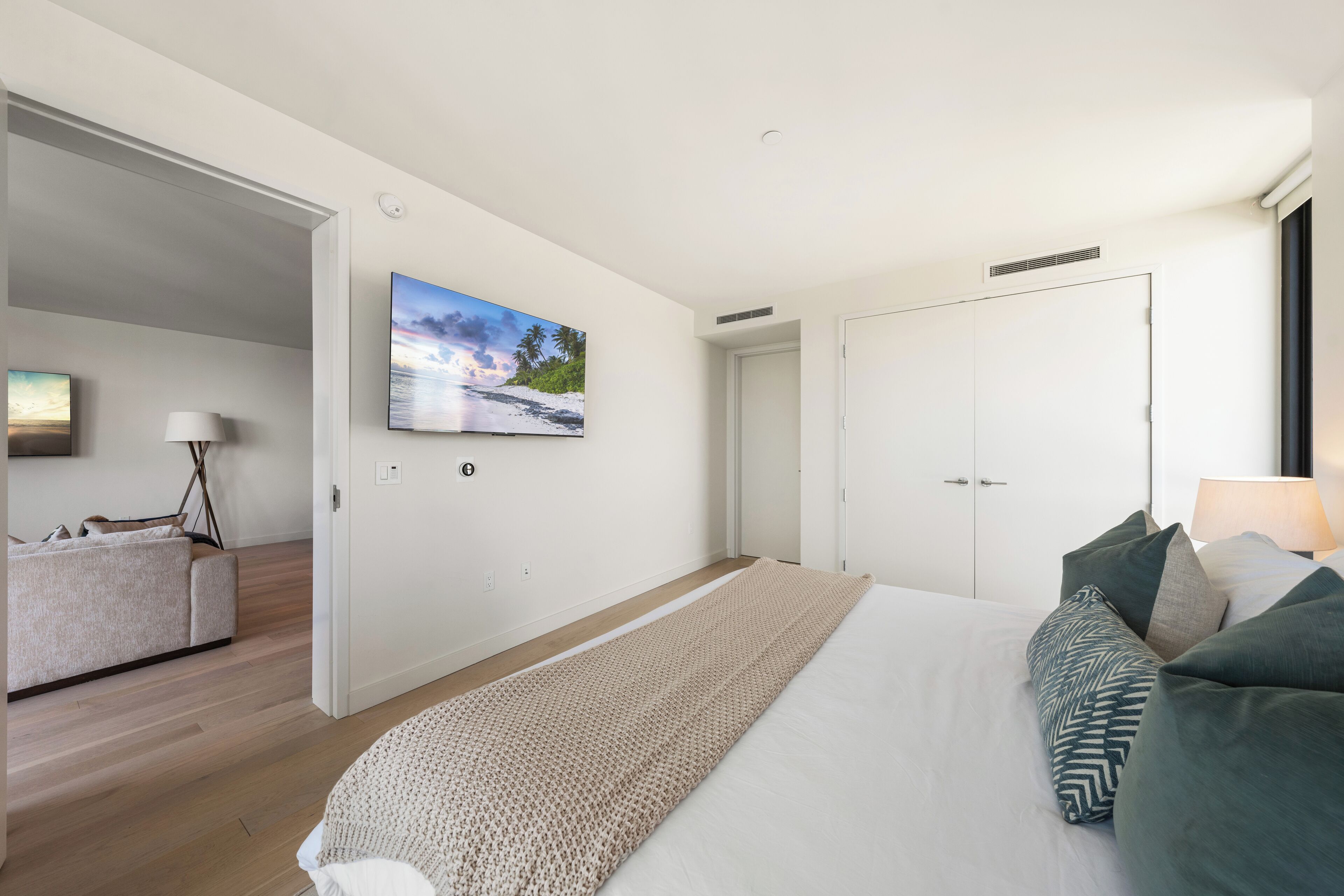 A modern bedroom with a large bed, a flat-screen TV on the wall, and a door leading to another room.