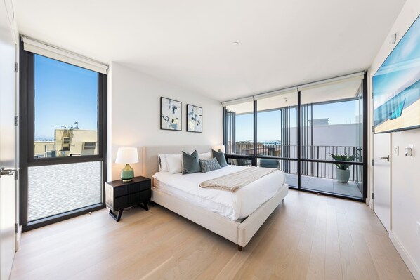 A modern bedroom with a large bed, a nightstand, a window with blinds, and a balcony with a view of the city.