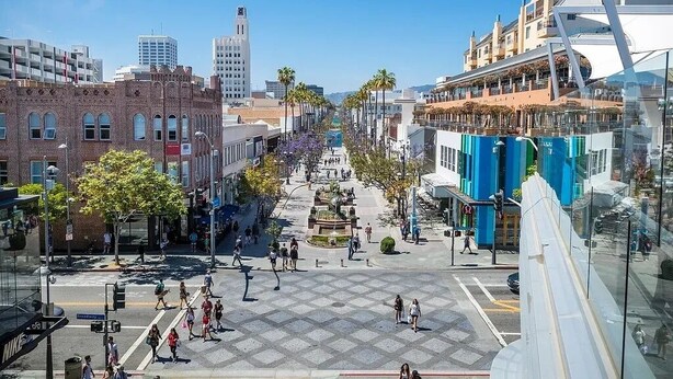 Una plaça urbana amb una font central, envoltada d'edificis moderns i històrics, i un cel serè.