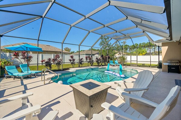 A covered outdoor swimming pool with lounge chairs, a table, and a grill. - 5BR Pool Retreat with Lanai, Fire Pit & Bikes (Palm Coast)