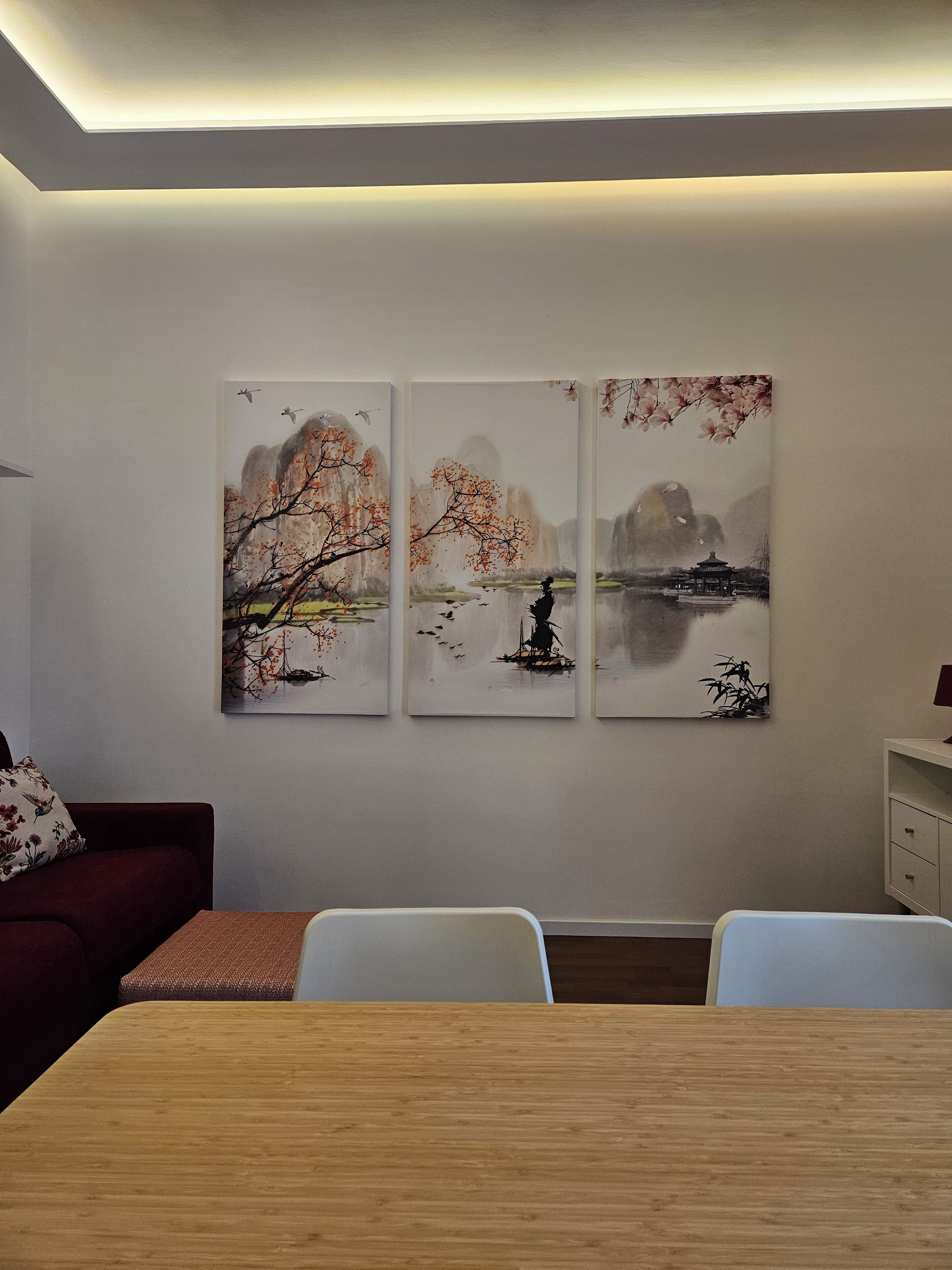 Un coin repas avec une table en bois, des chaises blanches et trois tableaux au mur représentant des paysages paisibles.