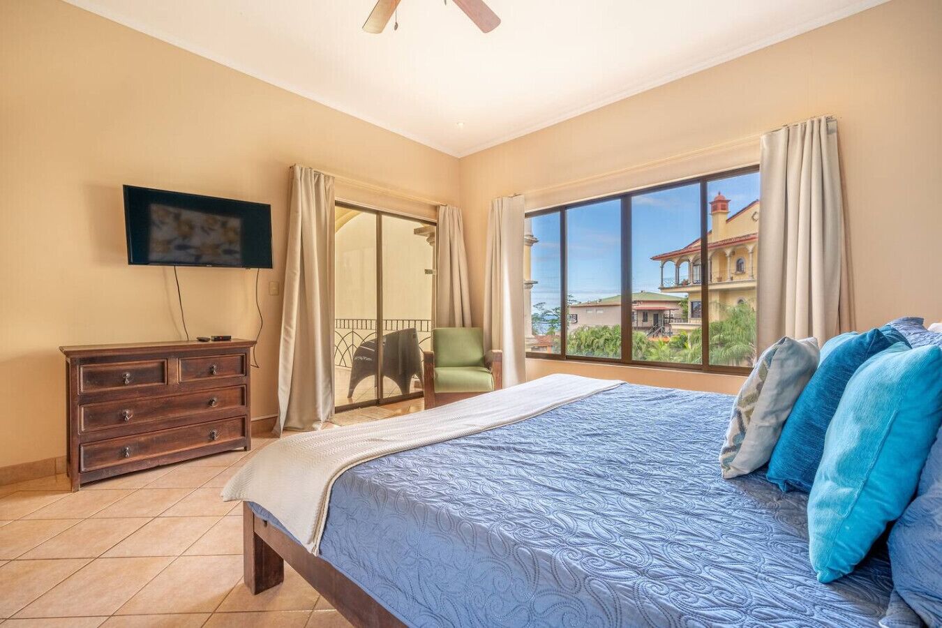 A bedroom with a large bed, a TV on the wall, a sliding glass door to a balcony, and a chair.