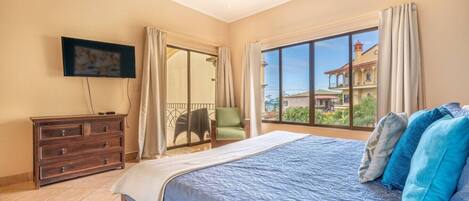 A bedroom with a large bed, a TV on the wall, a sliding glass door to a balcony, and a chair.