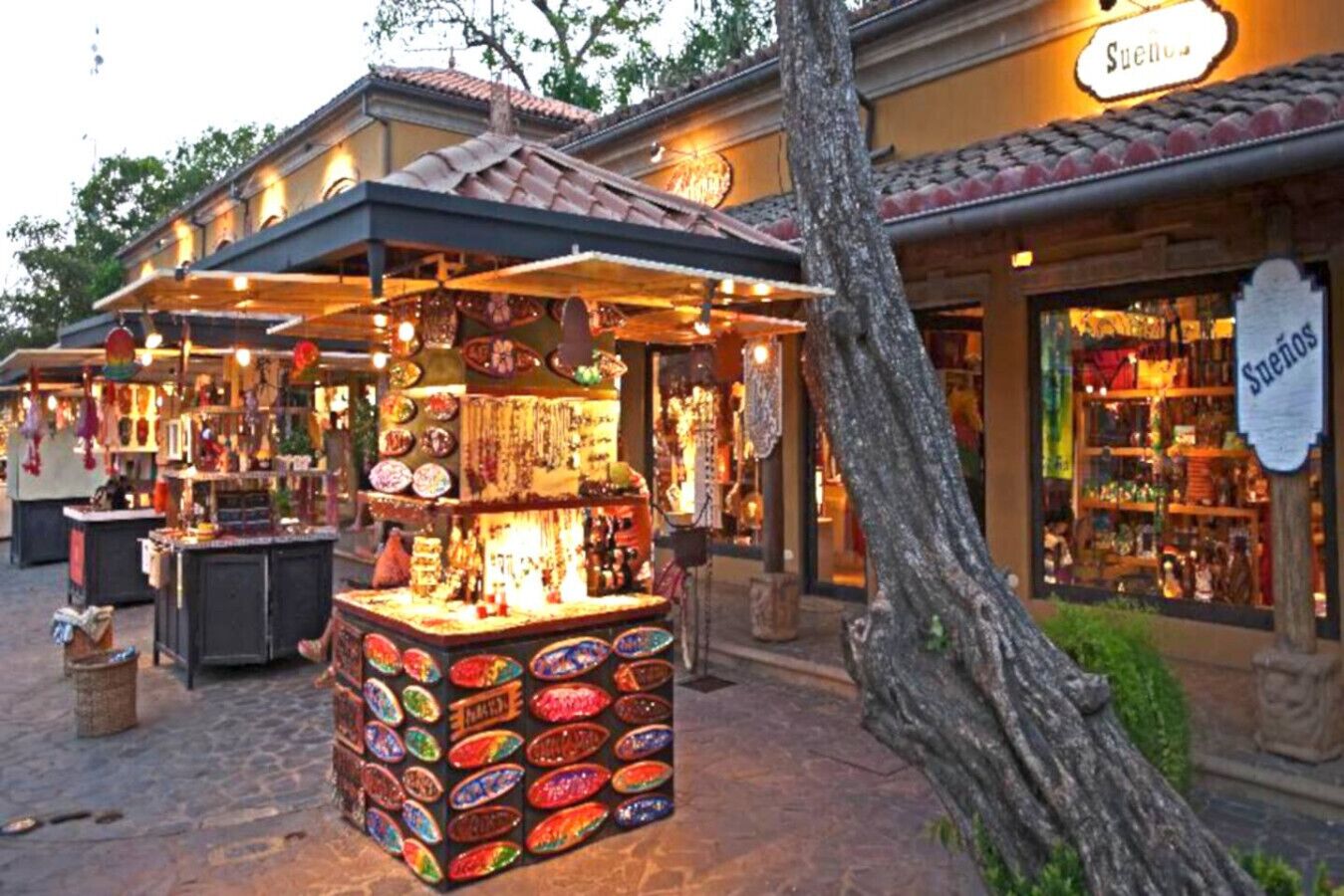 A street market with a central stall selling colorful items, surrounded by shops with visible signage.