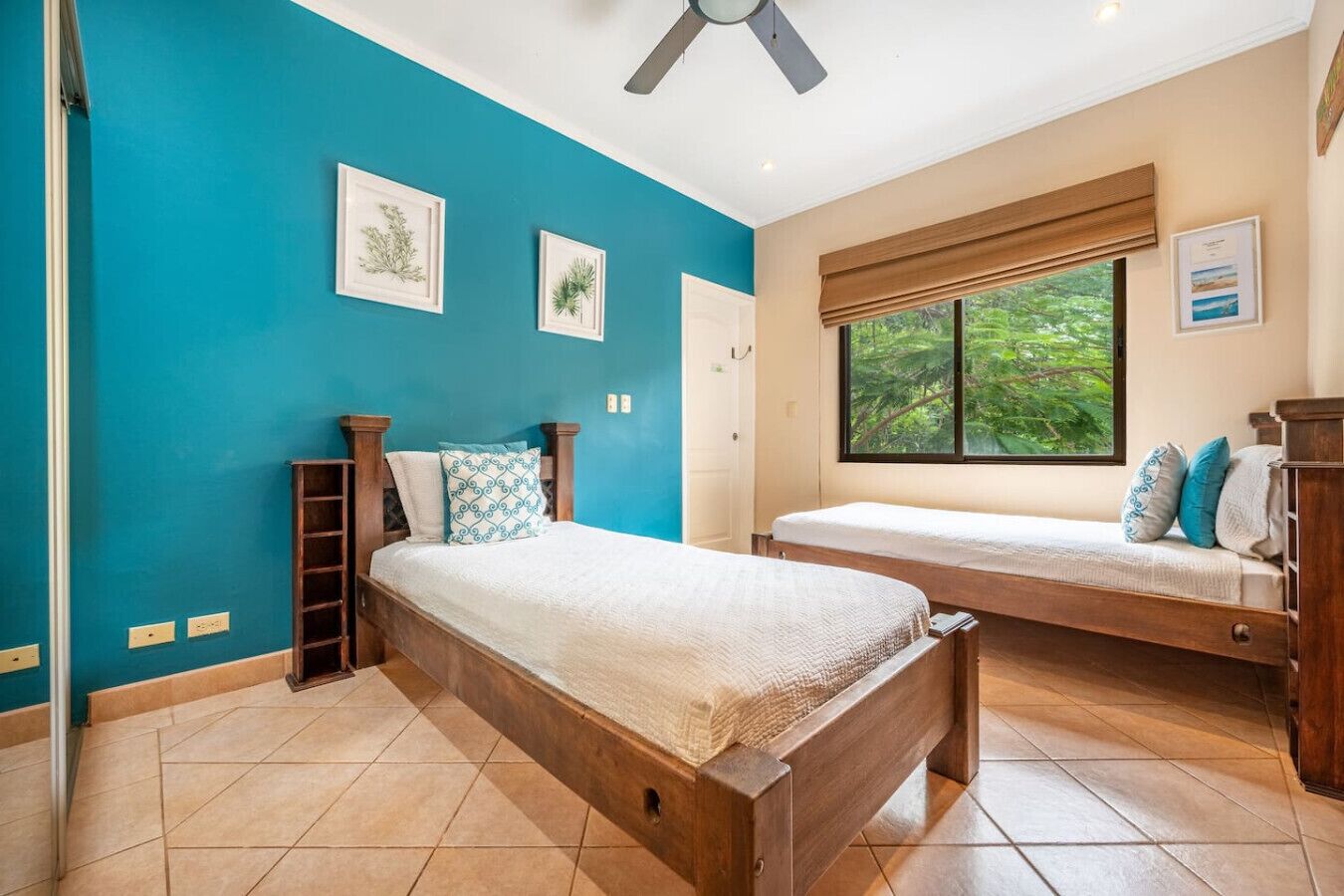 A bedroom with two beds, a ceiling fan, a window with a view of greenery, and framed pictures on the wall.