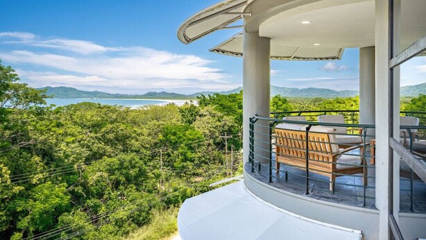 Ein modernes Gebäude mit einem Balkon, der auf eine üppige grüne Landschaft und den fernen Blick auf das Meer blickt.