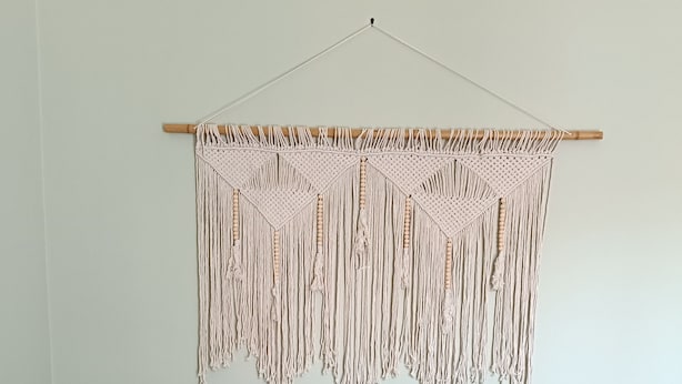 Een groene bank met drie kussens, een houten bijzettafeltje met een afstandsbediening, en een wanddecoratie van macramé.