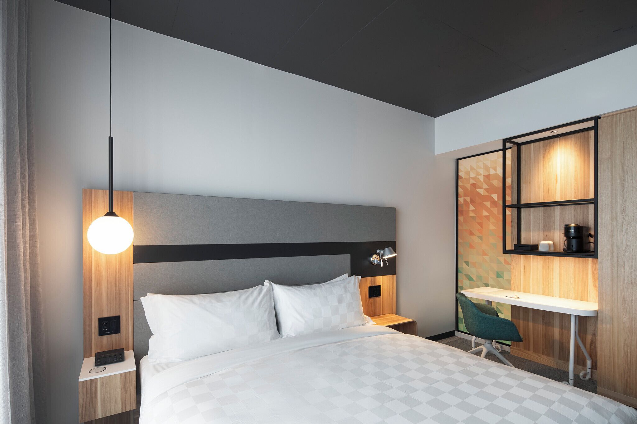 A modern hotel room with a bed, bedside table, lamp, desk, chair, and built-in shelving.
