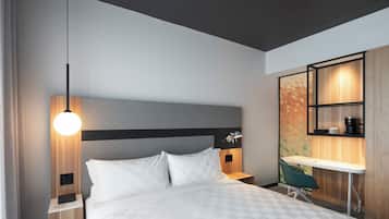 A modern hotel room with a bed, bedside table, lamp, desk, chair, and built-in shelving.