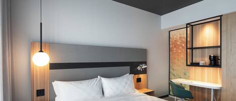 Une chambre d’hôtel moderne équipée d’un lit, d’une table de chevet, d’une lampe, d’un bureau, d’une chaise et d’étagères intégrées.