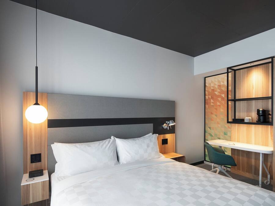 A modern hotel room with a bed, bedside table, lamp, desk, chair, and built-in shelving.