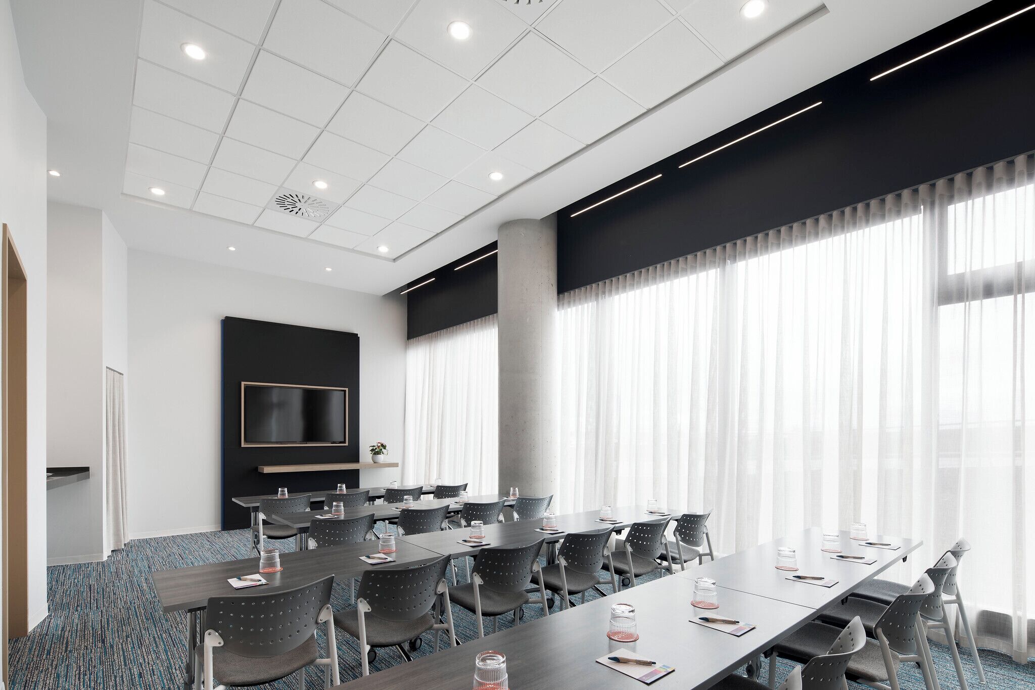 A conference room with a long table, chairs, a flat-screen TV, and large windows with sheer curtains.