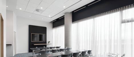 A conference room with a long table, chairs, a flat-screen TV, and large windows with sheer curtains.